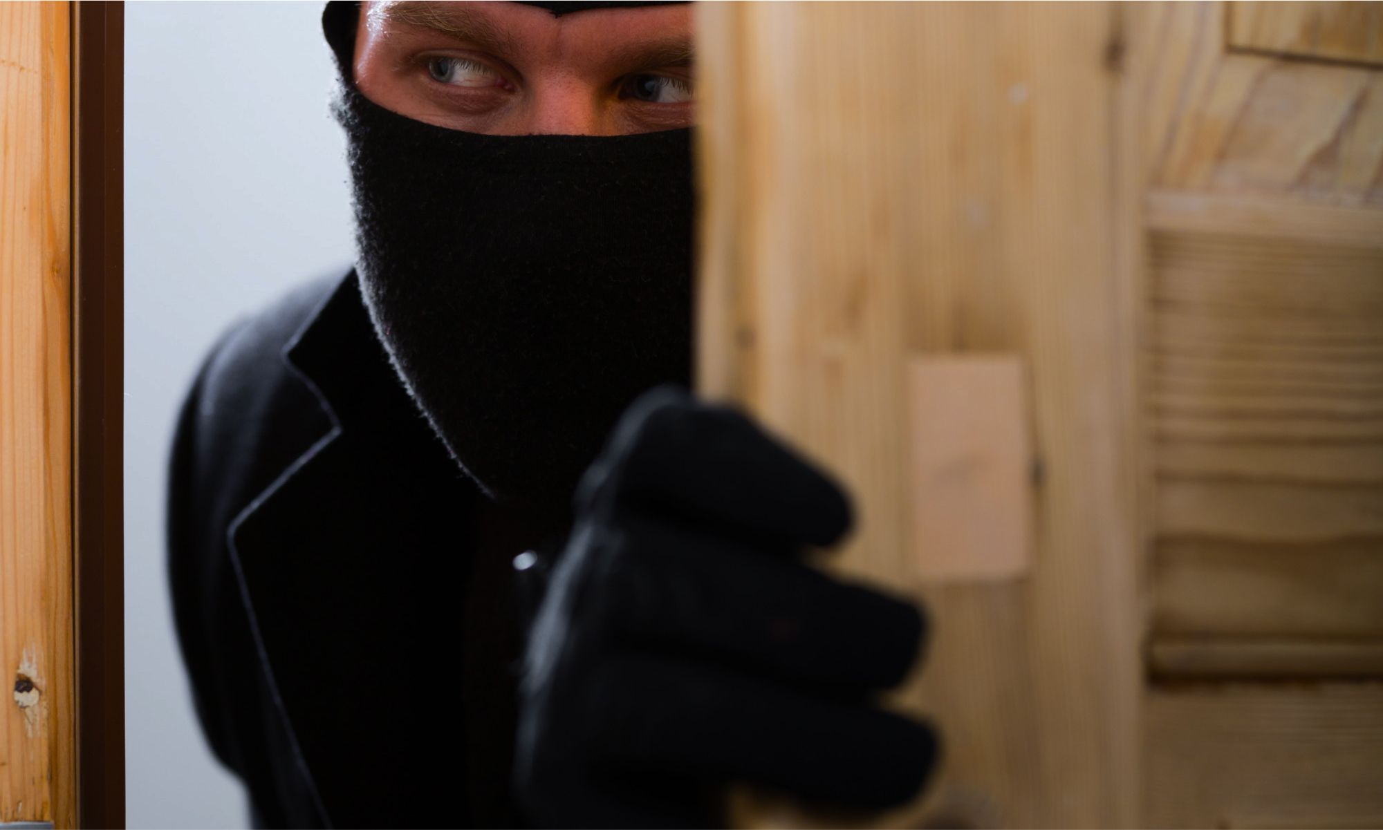 Home Invasions in U.S -Masked intruder in black peeking through a partially opened wooden door, appearing stealthy.
